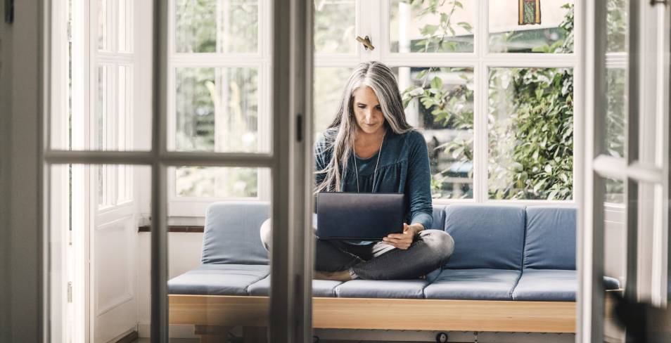 Older woman sitting cross-legged on a couch looking at her laptop.